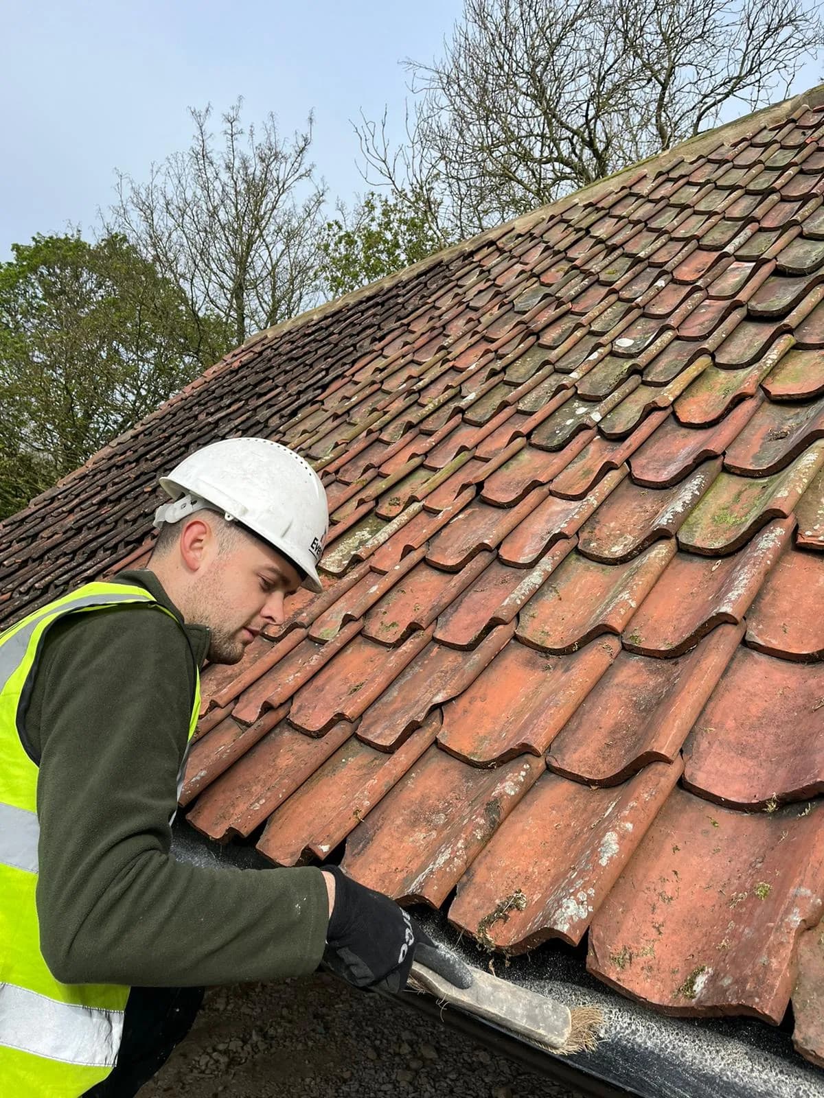 Evenii operative in branded hard hat and hi-vis working on the clay pantile barn roof, clearing the gutter line during the re-roof.