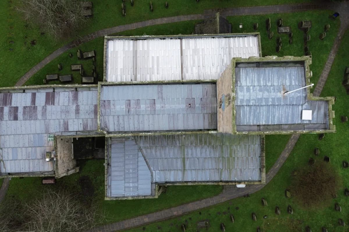 Wide aerial of stone church roof showing multiple lead-sheet bays, footprint context with pathways and graveyard — heritage roofing capability